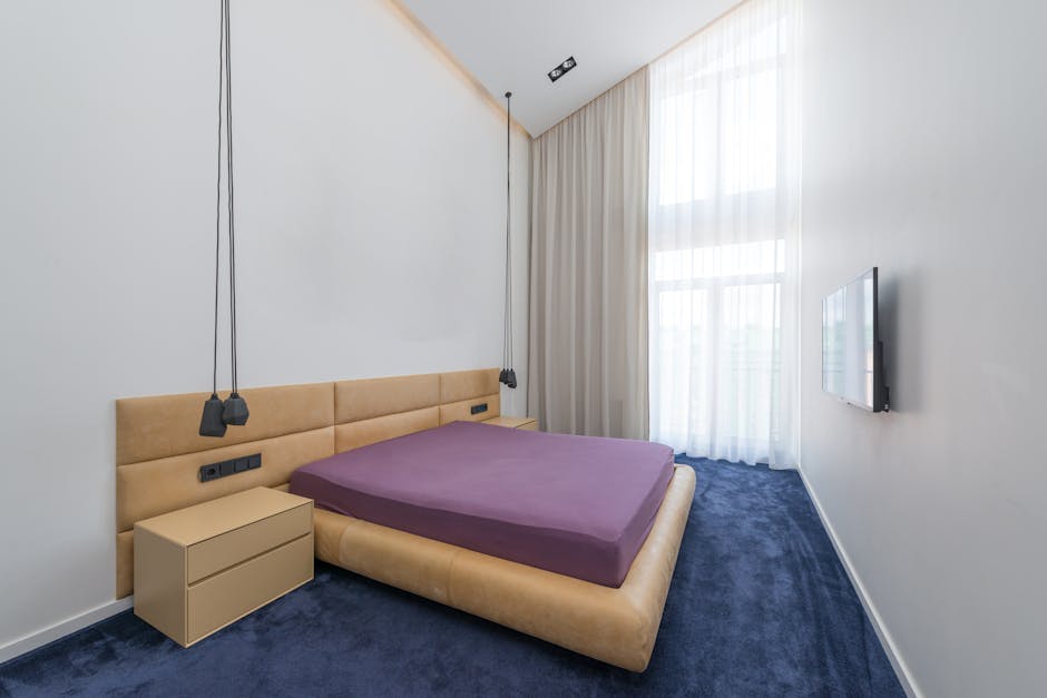 A modern bedroom featuring a beige upholstered bed with a purple mattress. The room has white walls, and a large window draped with sheer white curtains allowing natural light to fill the space. A small beige bedside table is positioned next to the bed, and black pendant lights hang from the ceiling above the headboard. The floor is covered with a dark blue carpet, and a flat-screen television is mounted on the wall opposite the bed. The room appears clean and well-maintained, suitable for surface cleaning, sanitisation, and general domestic cleaning services provided by Carpet Cleaning Marylebone.