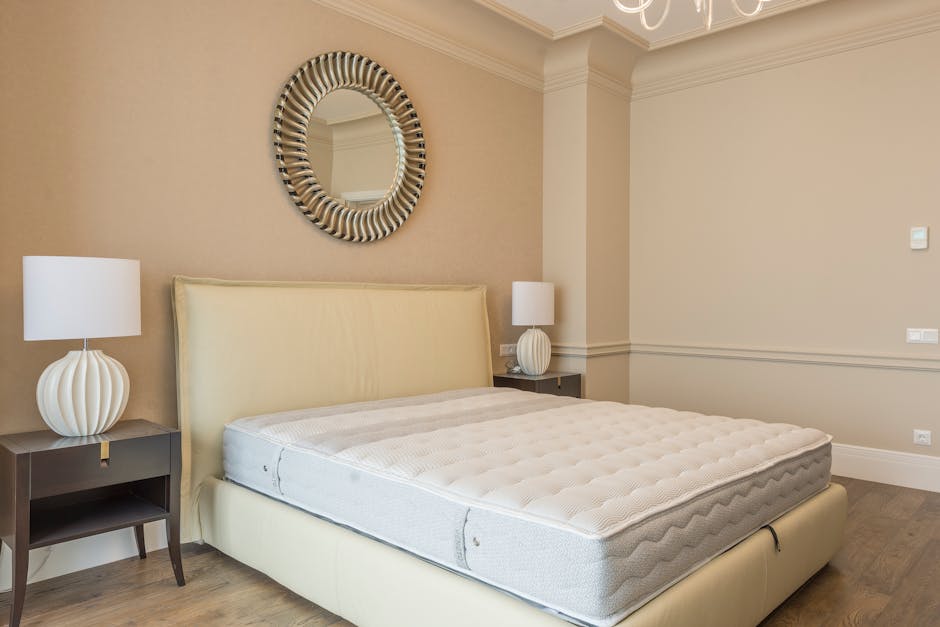 A modern bedroom featuring a neatly made double bed with a light beige upholstered headboard and a clean, white mattress. On each side of the bed, there are dark wooden nightstands, each topped with a white, ribbed ceramic table lamp. Above the bed, a round decorative mirror with a striped, textured frame hangs on the wall. The room has soft, neutral-colored walls and crown molding along the ceiling. The flooring appears to be wood or laminate, contributing to an overall tidy and hygienic appearance, suitable for surface cleaning and domestic sanitisation services provided by Carpet Cleaning Marylebone as referenced in the Gloucester Place mattress sanitising and Westminster rules context.