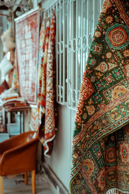 The image displays a selection of intricately patterned Persian or Oriental rugs hanging vertically on a metal rack in a well-lit room, possibly a rug cleaning shop such as Carpet Cleaning Marylebone. The rugs feature vibrant colors including reds, greens, and golds with detailed geometric and floral designs. The setting appears to be a commercial space with wooden flooring and a white wall with a patterned metal gate in the background. The rugs are clean and well-maintained, suggesting professional surface cleaning and deep cleaning processes aimed at preserving the intricate details and hygienic quality of the textiles. This arrangement showcases the quality of domestic and commercial rug sanitisation offered by Carpet Cleaning Marylebone, emphasizing their expertise in maintaining clean, hygienic surfaces through thorough cleaning procedures.
