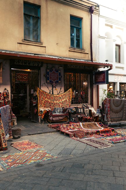 The image shows a street scene outside a shop with an open storefront displaying a variety of colorful Persian rugs and carpets laid out on the sidewalk and draped over furniture. The rugs feature intricate patterns and vibrant colors, with some rolled or partially folded. The shop interior and exterior walls are visible, with two upper windows and a beige facade. The scene is illuminated with natural daylight, highlighting the textures and details of the rugs, which appear clean and well-maintained. Signs of professional cleaning or surface maintenance are not visible, but the presentation suggests attention to hygiene and presentation typical of domestic or commercial surface cleaning. The presence of the rugs outside the shop emphasizes the importance of thorough surface cleaning and care in maintaining high standards of hygiene and appearance, aligned with services provided by Carpet Cleaning Marylebone, as referenced on the page about rug cleaning in Marylebone High Street.