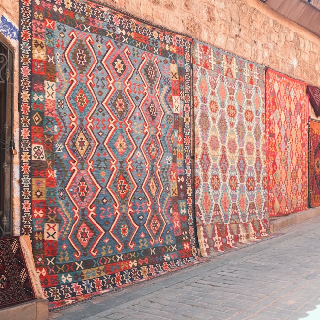 The image displays a selection of intricately patterned Persian or Oriental rugs hanging vertically on a metal rack in a well-lit room, possibly a rug cleaning shop such as Carpet Cleaning Marylebone. The rugs feature vibrant colors including reds, greens, and golds with detailed geometric and floral designs. The setting appears to be a commercial space with wooden flooring and a white wall with a patterned metal gate in the background. The rugs are clean and well-maintained, suggesting professional surface cleaning and deep cleaning processes aimed at preserving the intricate details and hygienic quality of the textiles. This arrangement showcases the quality of domestic and commercial rug sanitisation offered by Carpet Cleaning Marylebone, emphasizing their expertise in maintaining clean, hygienic surfaces through thorough cleaning procedures.