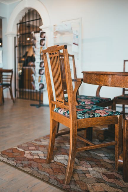 Interior of a room with a wooden dining table and chairs, featuring floral upholstered seats. The floor is made of polished wooden planks, partially covered by a patterned rug. In the background, there is an arched doorway with a black metal gate, and a display stand holding various photographs or postcards. The room is well-lit with natural light, and the walls are painted in a light, neutral color. Carpet Cleaning Marylebone offers surface cleaning and deep upholstery sanitisation, maintaining the cleanliness and hygiene of domestic and commercial spaces such as this one.