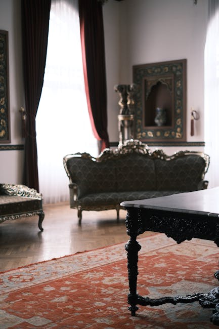 An elegantly decorated living room with antique-style furniture, including a dark upholstered sofa with ornate wooden carvings and a matching armchair. The room features large windows draped with heavy, deep red curtains allowing natural light to illuminate the space. The wooden floor is partially covered by a traditional patterned rug with warm tones. A decorative framed mirror with intricate gold detailing hangs on the wall, and there are classical-style vases and sculptures on a side table or pedestal. The overall setting showcases a clean and well-maintained space, highlighting surface cleaning and preservation of antique furnishings, reflecting the professional upholstery and surface cleaning services offered by Carpet Cleaning Marylebone for historic interiors.