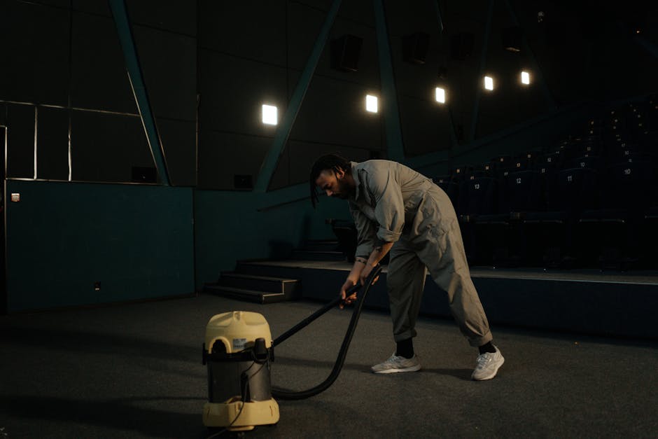 Close-up of a black vacuum cleaner head with a control switch, actively cleaning a patterned beige and green area rug with floral motifs, in a well-lit room. The vacuum is being used on a textured wov