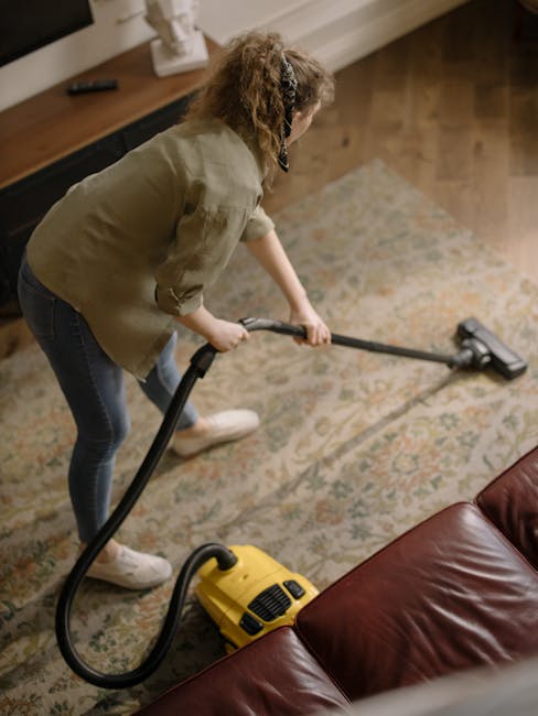 A woman using a yellow vacuum cleaner with a black hose to perform surface cleaning on a traditional patterned area rug in a living room. She is wearing a beige jacket, jeans, and white shoes, focused on deep cleaning the rug. The room has wooden flooring along the edges, a dark brown leather sofa in the foreground, and a wooden side table with decorative items and a TV in the background. The space is well-lit, highlighting the cleanliness and condition of the rug and surrounding surfaces, reflecting the professional domestic cleaning services offered by Carpet Cleaning Marylebone. The scene emphasizes thorough sanitisation and maintenance of textile surfaces within a home environment, aligning with the topic of spot hidden cleaning fees for Marylebone homeowners.