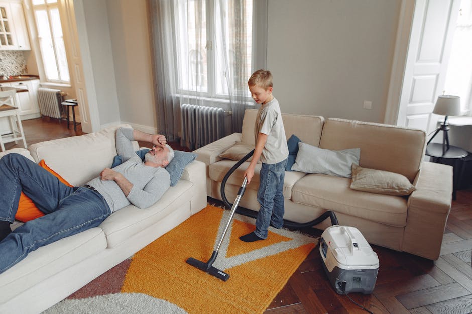 A young boy vacuuming a beige carpet in a bright living room with large windows, light-colored walls, and a white sofa. An older man with gray hair and a beard, wearing a gray sweater and jeans, is lying on the sofa, resting with his hand on his forehead. The room features a wooden floor, a yellow and white rug, cushions on the sofa, and a side table with a lamp. The vacuum cleaner operated by the boy is modern and white, with a black hose attached, and is positioned near the sofa on the wooden floor, which appears clean and well-maintained. The scene emphasizes domestic surface cleaning and deep cleaning processes in a residential setting, with natural lighting highlighting the tidy and hygienic environment, and is associated with professional cleaning services like those provided by Carpet Cleaning Marylebone.