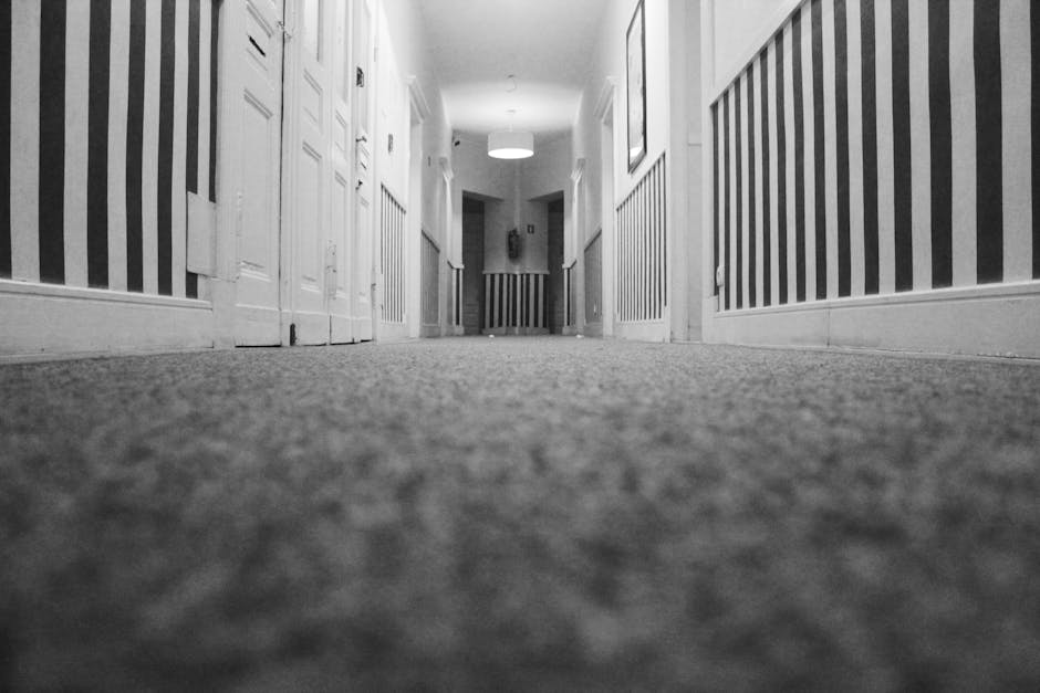 A low-angle view of a hallway with plush, patterned carpet flooring that appears clean and well-maintained, extending towards a distant doorway. The corridor is bordered by white wooden paneling and doors on either side, decorated with vertical black and white striped wallpaper. The ceiling features a modern light fixture emitting soft illumination, enhancing the sense of cleanliness and hygiene. The image suggests a thorough surface cleaning and maintenance suitable for residential or commercial spaces. For professional deep cleaning and sanitisation, [COMPANY_NAME] provides expert services aligned with tenancy-end carpet cleaning costs in Marylebone, as referenced on the page titled 'Tenancy-end carpet cleaning costs in Marylebone, Carpet Cleaning Marylebone'.