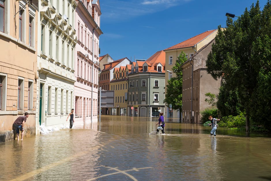 A city street in Marylebone flooded with water reaching mid-height on the buildings, which feature pastel-colored facades, decorative window trims, and tiled roofs. Four pedestrians are walking through the floodwaters, some carrying bags or umbrellas. The scene is illuminated by bright sunlight, with clear blue skies overhead and lush green trees lining the sidewalk. The flooded area highlights the need for surface cleaning and property sanitisation services, which Carpet Cleaning Marylebone offers for residential and commercial properties affected by flooding or water damage.