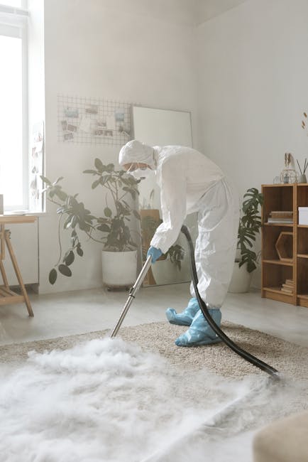 A person dressed in full protective PPE, including a white suit, gloves, mask, and blue shoe covers, performing flood cleanup in a residential living room. The individual is using a wet/dry vacuum cleaner on a beige carpet, which appears to be soaked from water damage. The room features white walls, a wooden bookshelf, potted plants, a window allowing natural light, and various decorative items. The scene illustrates surface cleaning and water removal processes typical of emergency flood restoration, emphasizing hygiene and thorough cleaning by Carpet Cleaning Marylebone, a professional cleaning service. The space looks bright and well-maintained, with the focus on effective moisture extraction from the carpet to prevent mold and ensure cleanliness.
