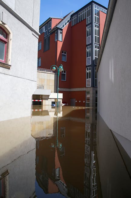 A city street in Marylebone flooded with water reaching mid-height on the buildings, which feature pastel-colored facades, decorative window trims, and tiled roofs. Four pedestrians are walking through the floodwaters, some carrying bags or umbrellas. The scene is illuminated by bright sunlight, with clear blue skies overhead and lush green trees lining the sidewalk. The flooded area highlights the need for surface cleaning and property sanitisation services, which Carpet Cleaning Marylebone offers for residential and commercial properties affected by flooding or water damage.